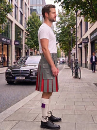 A man standing on a city sidewalk wearing a gray kilt with red pleated detailing, a white t-shirt, white socks, and black leather shoes, with a street scene in the background.