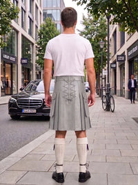 A man viewed from behind wearing a light gray kilt with lace-up back detailing, a white t-shirt, cream knee-high socks, and black shoes, standing on a city sidewalk.