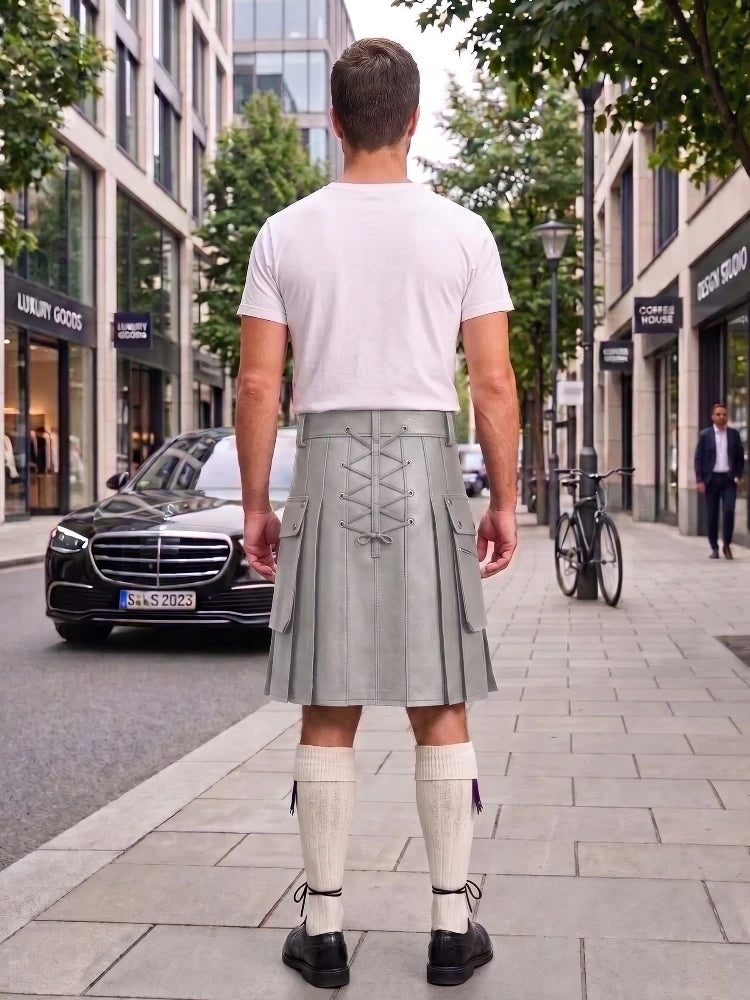 A man viewed from behind wearing a light gray kilt with lace-up back detailing, a white t-shirt, cream knee-high socks, and black shoes, standing on a city sidewalk.