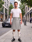 A man stands on a city sidewalk wearing a light gray pleated kilt with double-breasted buttons, a white t-shirt, white knee-high socks, and black leather shoes. A black Mercedes-Benz car and urban buildings are visible in the background.