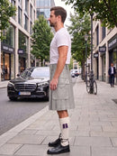 A man standing on a city sidewalk wearing a light gray pleated kilt, white t-shirt, white knee-high socks with purple accents, and black leather shoes. A black Mercedes-Benz car and urban buildings are visible in the background.