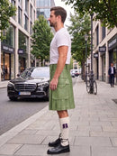 A man standing on a city sidewalk wearing a light olive green leather kilt, a white t-shirt, white socks, and black leather shoes. The background shows a street with a black Mercedes-Benz car, storefronts, and trees.