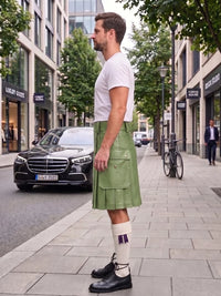 A man standing on a city sidewalk wearing a light olive green leather kilt, a white t-shirt, white socks, and black leather shoes. The background shows a street with a black Mercedes-Benz car, storefronts, and trees.
