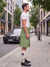 A man standing on a city sidewalk wearing a white crew-neck t-shirt, a pleated sage green kilt, white socks with purple stripes, and black leather oxford shoes. A black Mercedes-Benz car and a bicycle are parked on the street behind him.