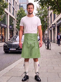 A man stands on a city sidewalk wearing a light green leather kilt, a white t-shirt, cream knee-high socks, and black leather shoes. The background shows a blurred street scene with buildings, a black Mercedes-Benz, and a bicycle.