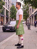 A man standing on a city sidewalk wearing a light green kilt, white t-shirt, white knee-high socks with purple accents, and black leather shoes. A black Mercedes-Benz car is parked on the street behind him, with trees and storefronts in the background.