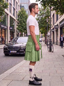 A man standing on a city sidewalk wearing a light green pleated kilt, a white short-sleeve t-shirt, white socks, and black leather shoes. A black Mercedes-Benz car and a bicycle are visible in the background on a city street.