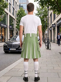 A man stands on a city sidewalk facing away from the camera, wearing a white short-sleeve T-shirt and a light green pleated kilt with white vertical stripe details. He is also wearing white knee-high socks and black shoes.