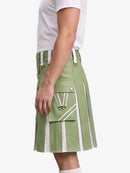 A side view of a model wearing a sage green kilt with white vertical stripes, silver buckles, and a large front cargo pocket with a white 'W' logo, paired with a white t-shirt against a light gray background.