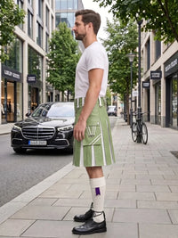 A man standing on a city sidewalk wearing a light green kilt with white vertical stripes, a white t-shirt, white socks, and black leather shoes. A black Mercedes-Benz car and a bicycle are parked on the street behind him.