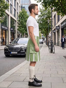 A man stands on a city sidewalk wearing a white crew-neck t-shirt and a light green pleated kilt with white vertical stripe details, paired with cream knee-high socks and black leather shoes. A black Mercedes-Benz car and a bicycle are visible in the background of the urban street scene.