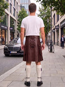 A man standing on a city sidewalk, viewed from behind, wearing a dark brown leather kilt with lace-up detailing, a white t-shirt, white knee-high socks, and black shoes. Buildings, trees, a black Mercedes-Benz, and a bicycle are visible in the background.