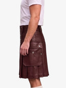 A man wearing a dark brown faux-leather kilt with multiple flap pockets and silver snap buttons, paired with a white t-shirt, shown from a side profile against a light gray background.