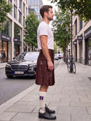 A man standing on a city sidewalk wearing a dark brown leather kilt, a white t-shirt, white socks with purple stripes, and black leather shoes. A black Mercedes-Benz car is parked on the street behind him, with trees and storefronts in the background.