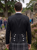 A man viewed from behind wearing a black formal jacket with silver-toned decorative buttons and a gray plaid kilt, standing outdoors in front of stone ruins and trees.