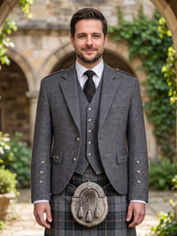A man wearing a traditional Scottish formal outfit with a gray three-piece jacket and waistcoat, a plaid kilt, a black tie, and a sporran, standing in front of a stone archway with green ivy.