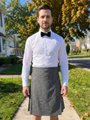 A man standing outdoors on a sidewalk wearing a white long-sleeve dress shirt, black bow tie, and a gray pleated kilt.