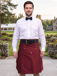 A man wearing a burgundy double-breasted kilt with gold buttons, a black belt, a white dress shirt, and a black bow tie, standing outdoors in front of a stone building and green lawn.