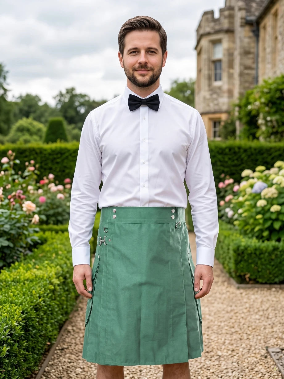 A man wearing a white dress shirt, black bow tie, and a green kilt stands in a garden setting with hedges and a stone building in the background.