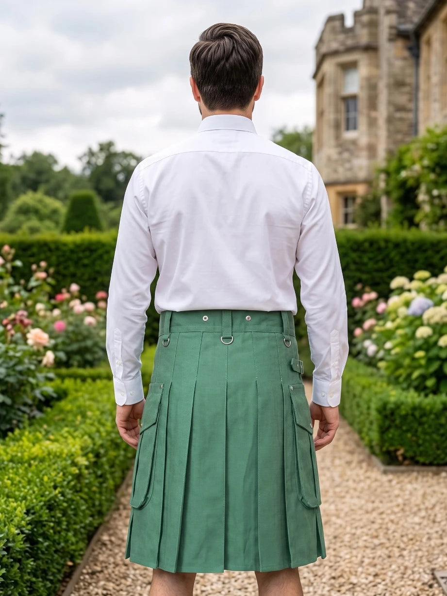 A man viewed from behind wearing a white long-sleeve dress shirt and a green pleated kilt, standing on a garden path with hedges and a stone building in the background.