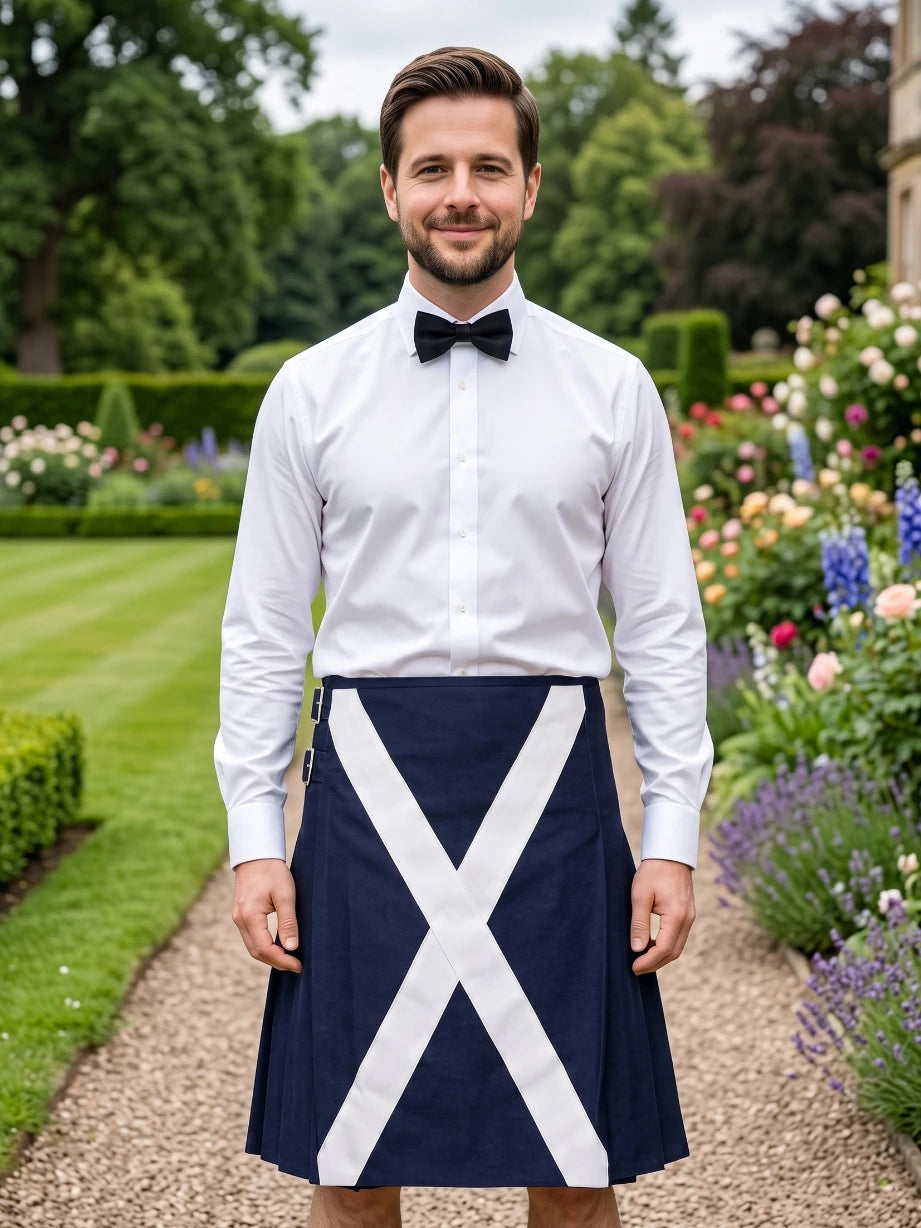 A man standing outdoors in a garden wearing a white long-sleeve dress shirt, black bow tie, and a navy blue kilt with a large white diagonal cross.