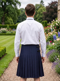 A man viewed from behind wearing a white long-sleeve dress shirt tucked into a dark navy pleated kilt with belt loops, standing on a garden path.