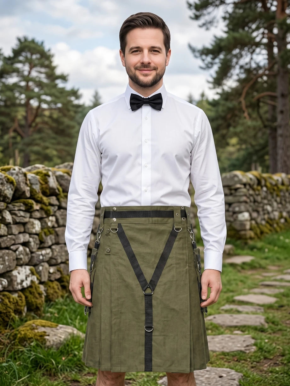 A man wearing a white dress shirt, black bow tie, and an olive green kilt with black suspenders, standing outdoors in front of a stone wall and trees.