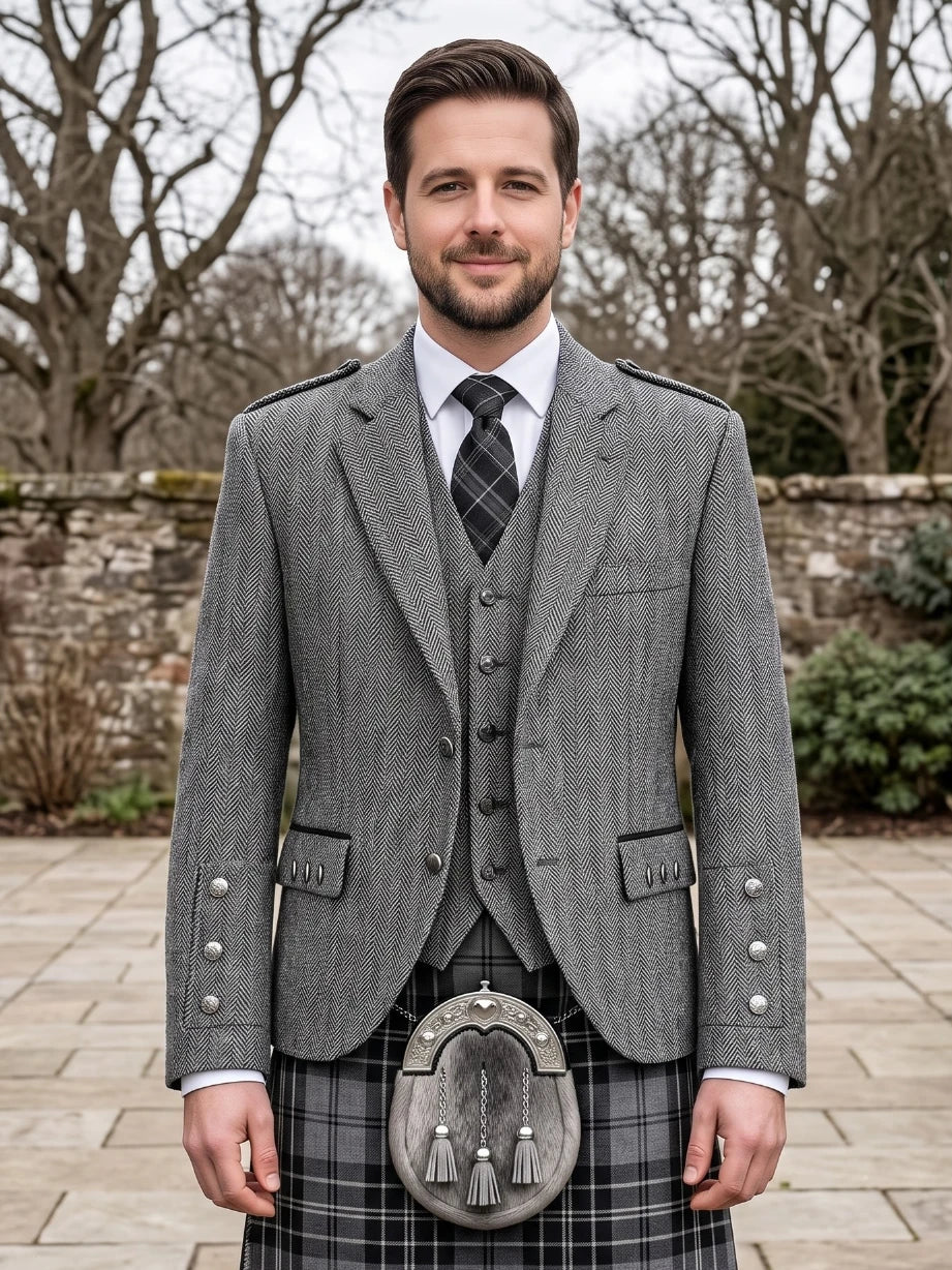 A man wearing a traditional Scottish formal outfit with a gray herringbone blazer, matching waistcoat, white shirt, patterned tie, and a gray plaid kilt with a sporran, standing outdoors in front of a stone wall and bare trees.