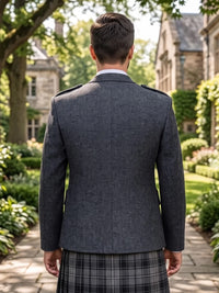 A man viewed from behind wearing a dark gray textured blazer with shoulder epaulets and a black and gray plaid kilt, standing on a stone path in a garden courtyard.