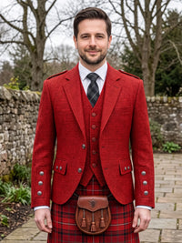 A man wearing a traditional Scottish formal outfit, featuring a textured red tweed blazer, matching red tweed waistcoat, a red and black plaid kilt, a brown leather sporran, and a dark patterned tie, standing outdoors in front of a stone wall.