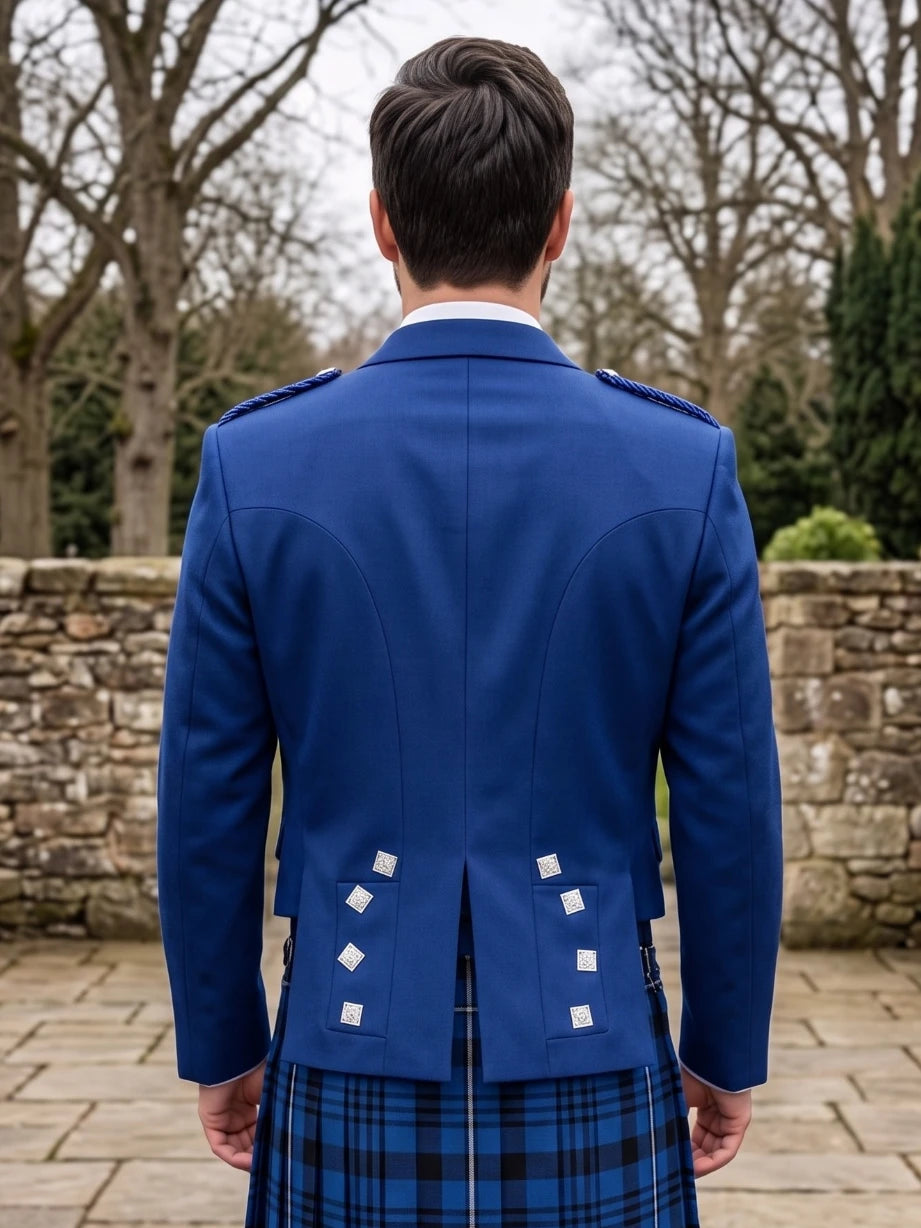 A man viewed from behind wearing a royal blue Scottish kilt jacket with white decorative buttons and a blue and black plaid kilt, standing outdoors in front of a stone wall and bare trees.