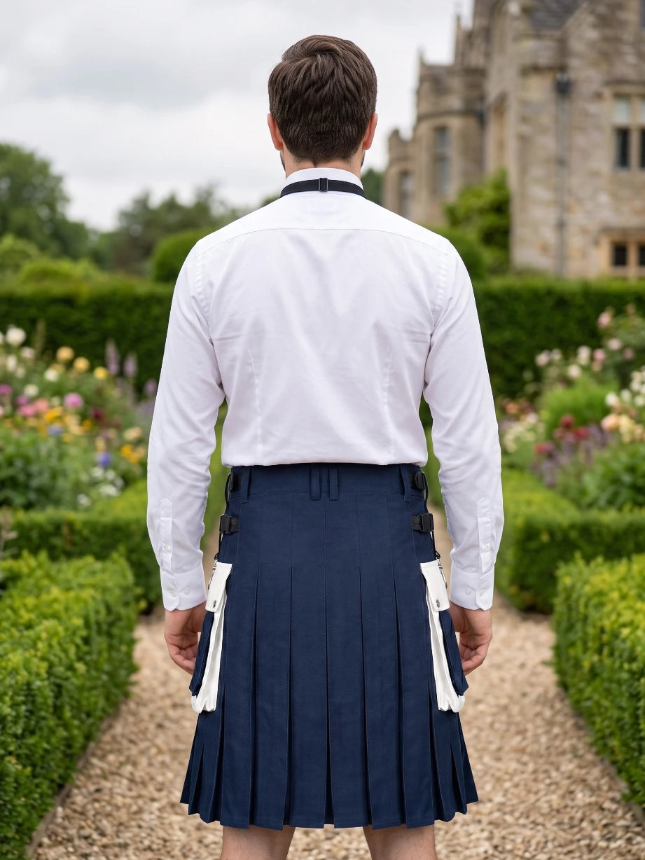 A man viewed from behind wearing a white long-sleeve dress shirt and a navy blue kilt with white wing panels, standing on a garden path with hedges and a stone building in the background.
