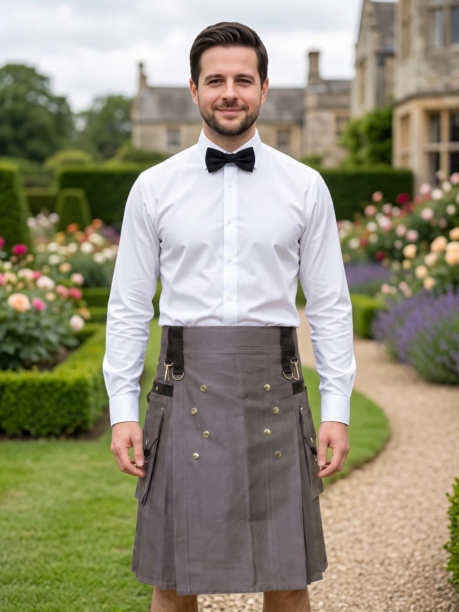 A man wearing a white dress shirt, black bow tie, and a gray kilt with black suspenders and brass buttons, standing in a garden path with a stone building in the background.