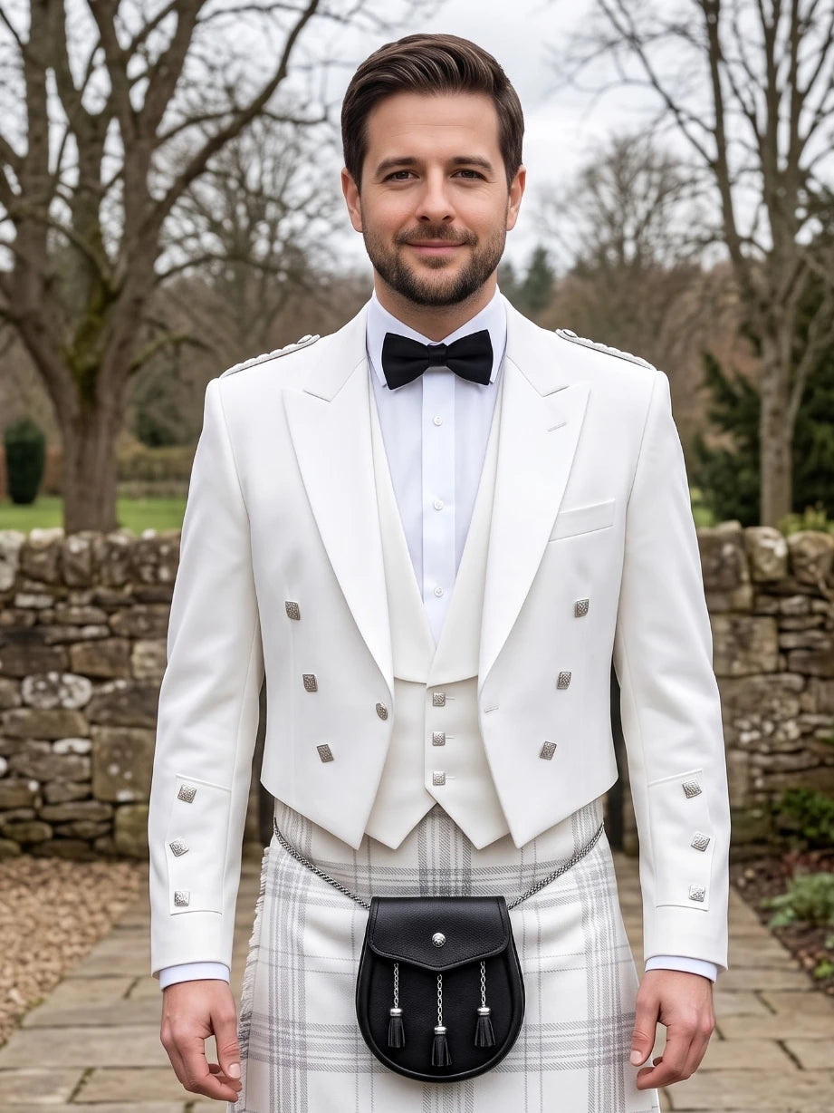 A man wearing a formal white double-breasted jacket with silver buttons, a white waistcoat, a white shirt, a black bow tie, and a black sporran on a white plaid kilt, standing outdoors in front of a stone wall and bare trees.