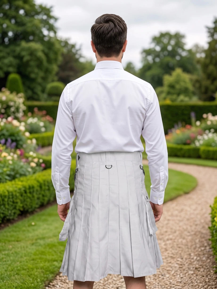 A man viewed from behind wearing a white long-sleeve dress shirt and a pleated light gray kilt, standing on a gravel path in a lush green garden.