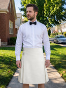 A man wearing a white long-sleeve dress shirt with vertical pleats, a black bow tie, and a cream-colored pleated kilt, standing on a suburban sidewalk.