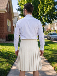 A man viewed from behind wearing a white long-sleeve button-down shirt and a cream pleated kilt, standing on a suburban sidewalk with houses and trees in the background.