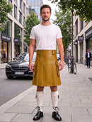 A man stands on a city sidewalk wearing a mustard yellow leather kilt over a white t-shirt, white knee-high socks, and black leather shoes. A black Mercedes-Benz car and a bicycle are visible in the background of the street scene.