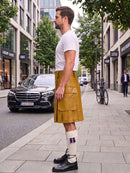 A man standing on a city sidewalk wearing a mustard yellow leather kilt, a white t-shirt, white socks, and black leather shoes. The background shows a street with a black Mercedes-Benz, trees, and buildings.