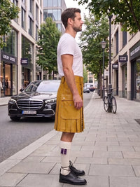 A man standing on a city sidewalk wearing a mustard yellow kilt, white t-shirt, white socks with purple stripes, and black leather shoes, with a street scene in the background.