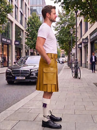 A man standing on a city sidewalk wearing a tan pleated kilt with a white t-shirt, white socks, and black leather shoes. The background shows a street with buildings, trees, and parked cars.