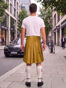 A man standing on a city sidewalk, viewed from behind, wearing a white t-shirt and a mustard yellow pleated leather kilt with white knee-high socks and black shoes.