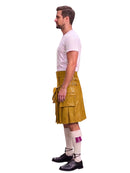 A man shown in profile wearing a mustard yellow leather kilt with multiple utility pockets and straps, a light pink t-shirt, white knee-high socks with a purple accent, and black leather shoes, isolated on a white background.