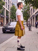 A man standing on a city sidewalk wearing a mustard yellow leather kilt, a white t-shirt, white knee-high socks, and black leather shoes. A black Mercedes-Benz car is parked on the street behind him, with trees and buildings in the background.