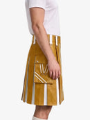 A mustard yellow faux-leather kilt with white vertical stripes and white strap details, shown on a model wearing a white t-shirt against a light gray background.