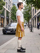 A man stands on a city sidewalk wearing a mustard yellow faux-leather kilt with white vertical stripes, a white t-shirt, cream knee-high socks, and black leather shoes. A black Mercedes-Benz car and a bicycle are visible in the background on a tree-lined street.