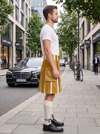 A man stands on a city sidewalk wearing a mustard yellow faux-leather kilt with white vertical stripes, a white t-shirt, cream knee-high socks, and black leather shoes. A black Mercedes-Benz car and a bicycle are visible in the background on a tree-lined street.