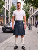 A man stands on a city sidewalk wearing a white crew-neck t-shirt and a dark navy blue pleated leather kilt with silver metal buttons. He is also wearing white knee-high socks and black leather shoes.