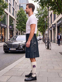 A man standing on a city sidewalk wearing a white crew-neck t-shirt, a dark navy blue leather kilt, white socks with a purple bow, and black leather shoes. A black Mercedes-Benz car is parked on the street behind him.