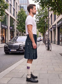A man standing on a city sidewalk wearing a dark green kilt, white t-shirt, white socks, and black leather shoes, with a street and buildings in the background.